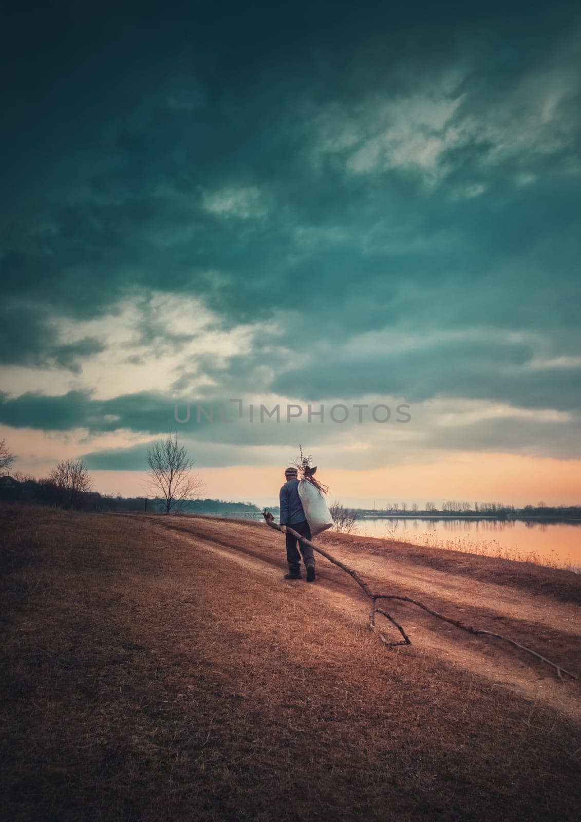 Rural evening scene, vertical photo with a lone nomad man carrying firewood and a bagful on his shoulder, going home walking a country dirt road near the lake. Moody and dramatic view.