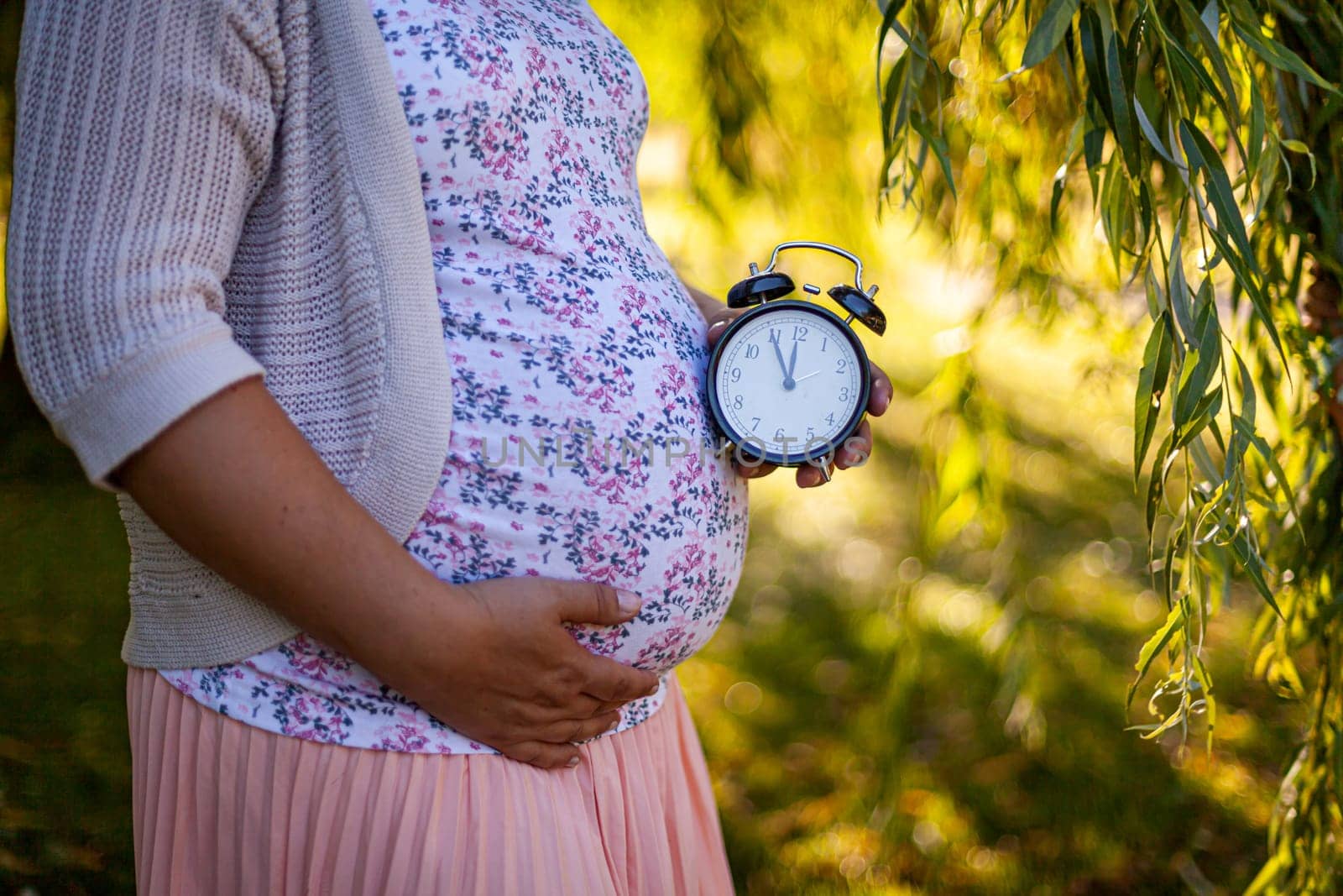 A pregnant women in a white dress in park holding a clock. High quality photo