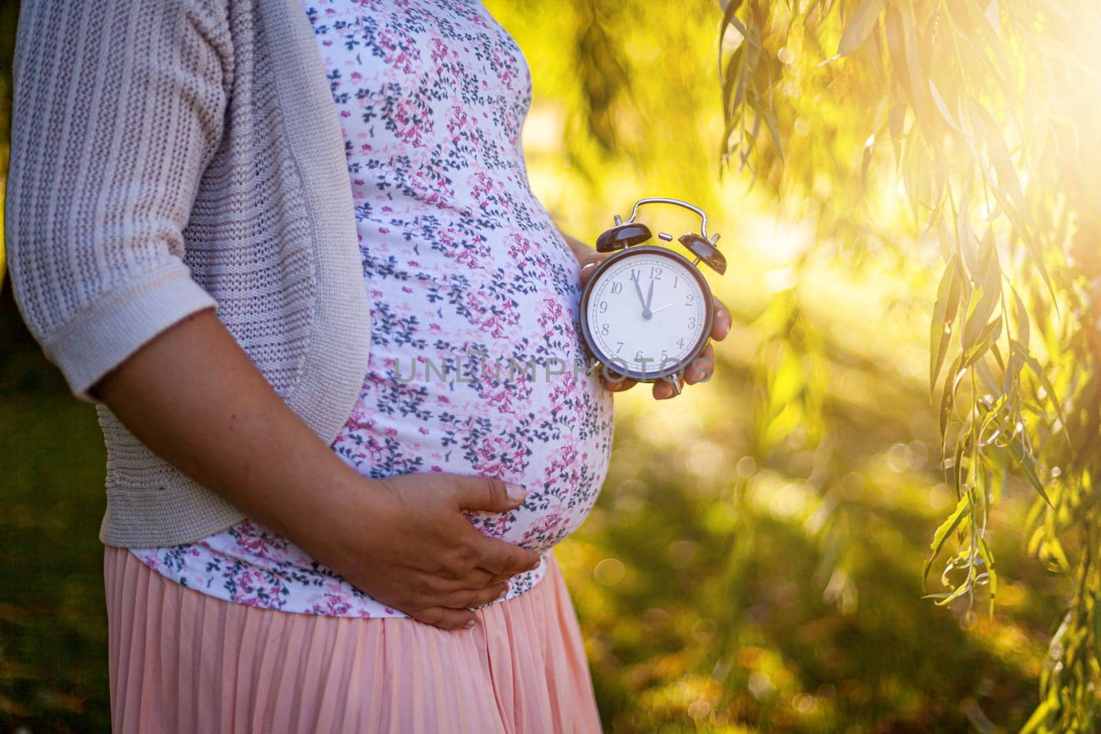 A pregnant women in a white dress in park holding a clock. High quality photo