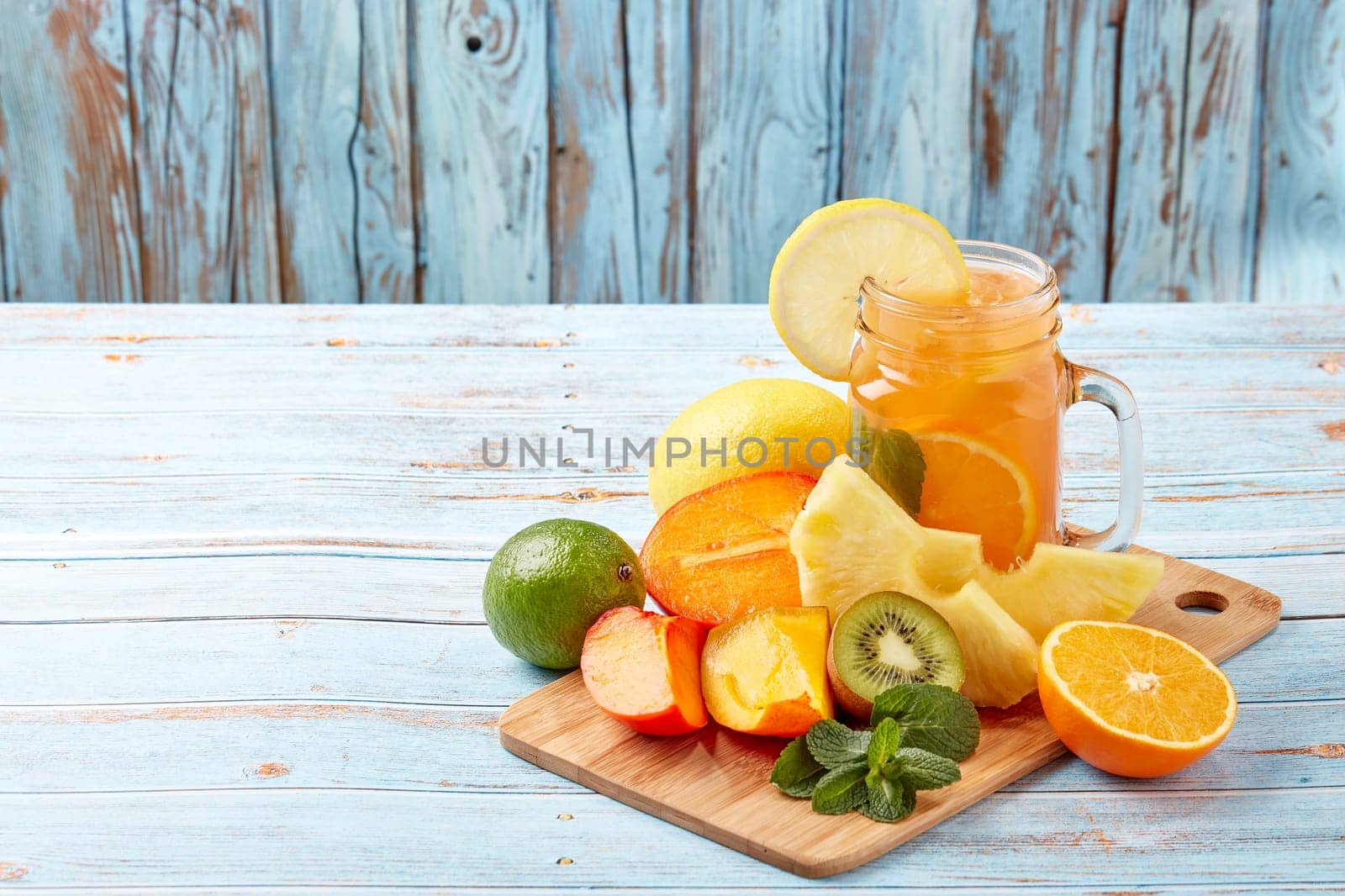 Fruit lemonade with ice, leaves of mint, lemon, pineapple, mango and lime slices standing on blue wooden table. Homemade lemonade with mix fruits. Healthy cool beverage, fruit juice