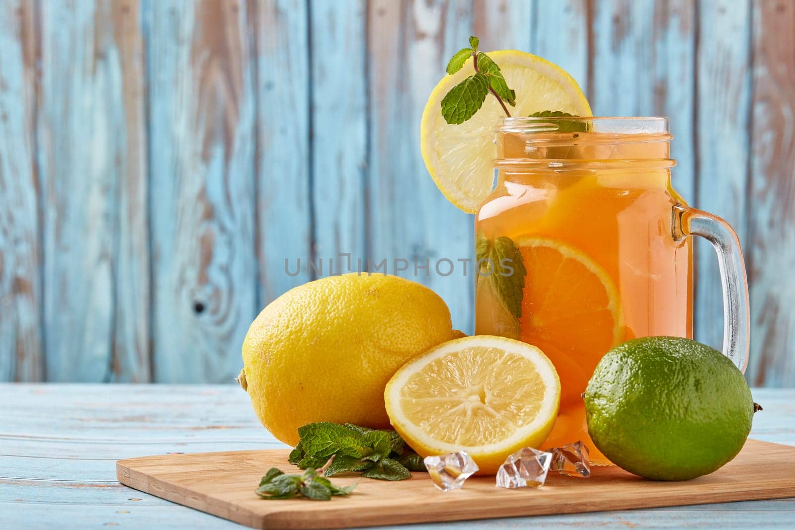 Citrus lemonade with ice, leaves of mint, lemon and lime slices standing on blue wooden table. Homemade lemonade with citruses. Healthy cool beverage
