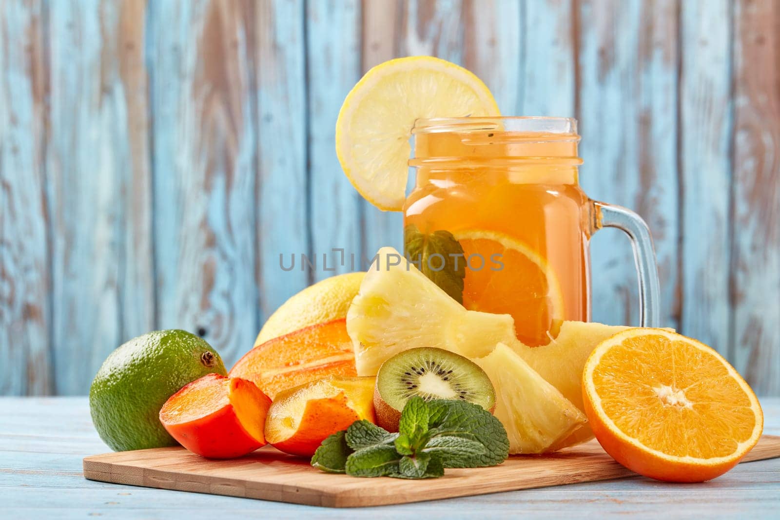 Fruit lemonade with ice, leaves of mint, lemon, pineapple, mango and lime slices standing on blue wooden table. Homemade lemonade with mix fruits. Healthy cool beverage, fruit juice