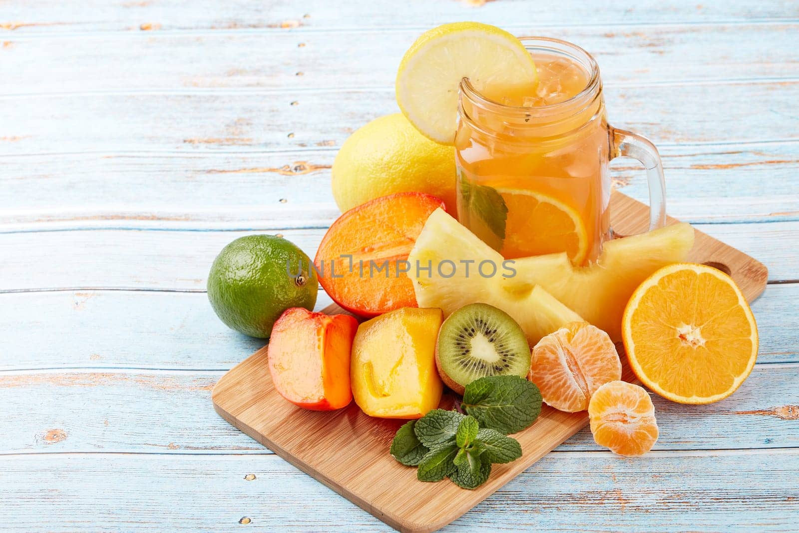 Fruit lemonade with ice, leaves of mint, lemon, pineapple, mango and lime slices standing on blue wooden table. Homemade lemonade with mix fruits. Healthy cool beverage, fruit juice