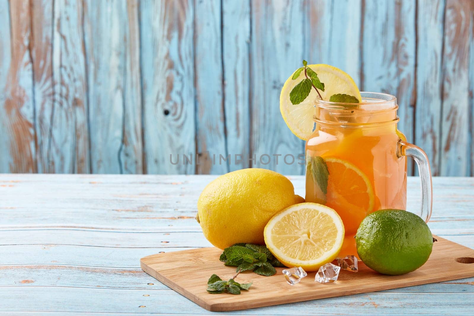 Citrus lemonade with ice, leaves of mint, lemon and lime slices standing on blue wooden table. Homemade lemonade with citruses. Healthy cool beverage