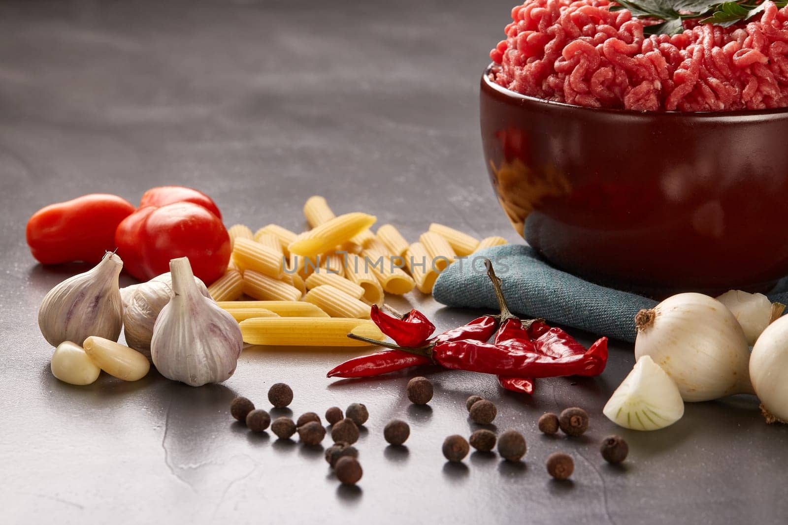 Still life with fresh forcemeat from beef in a bowl, vegetables, pasta and spices on a dark background.