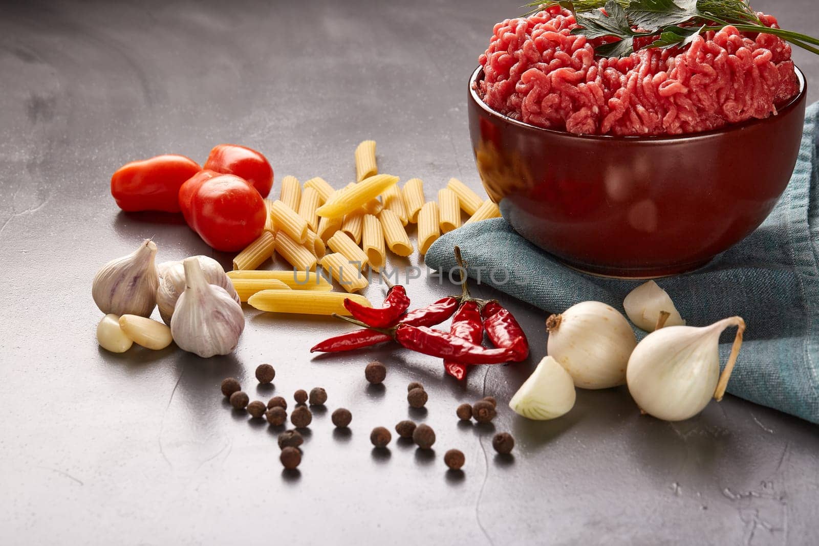 Still life with fresh forcemeat from beef in a bowl, vegetables, pasta and spices on a dark background.