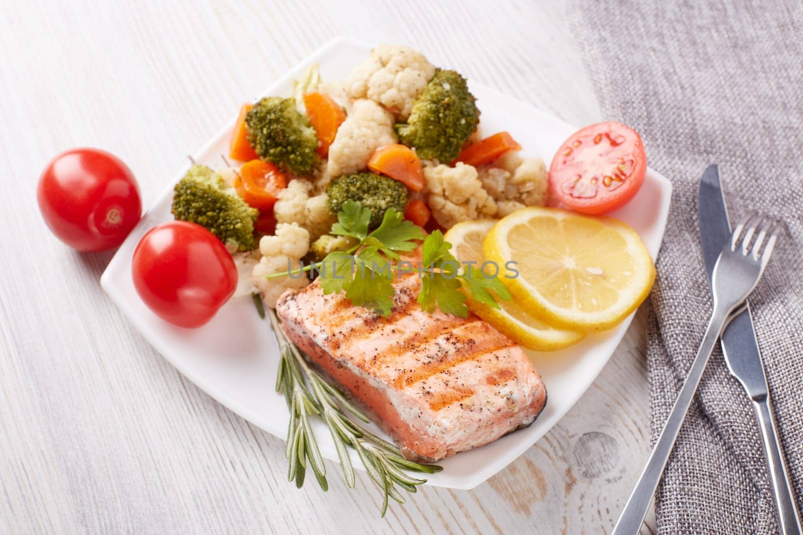 Grilled salmon fillet  garnished with vegetables, lemon, tomato, herbs lying on plate on wooden background. Top view.Healthy food. Keto diet.