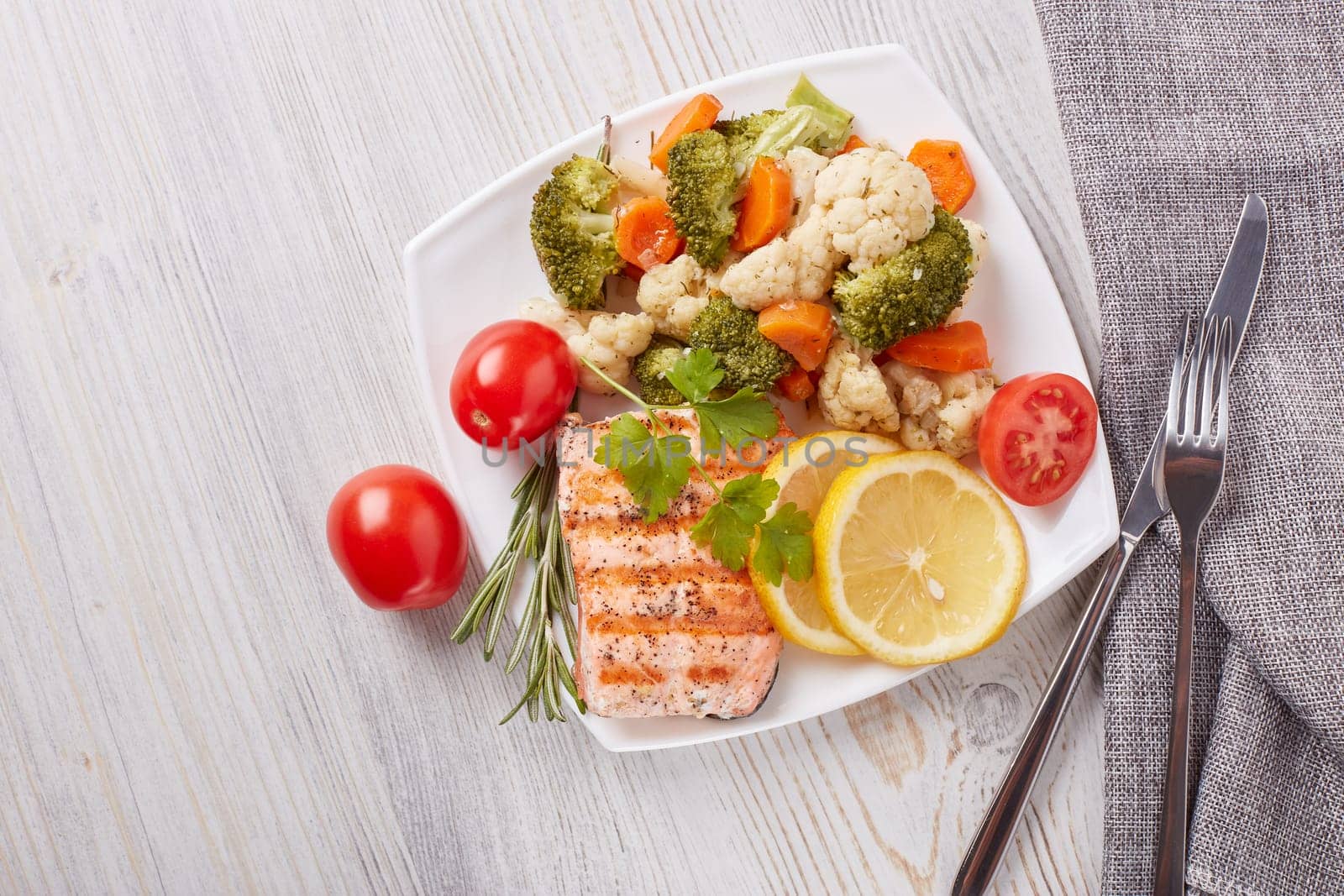 Grilled salmon fillet  garnished with vegetables, lemon, tomato, herbs lying on plate on wooden background. Top view with copy space.Healthy food. Keto diet.
