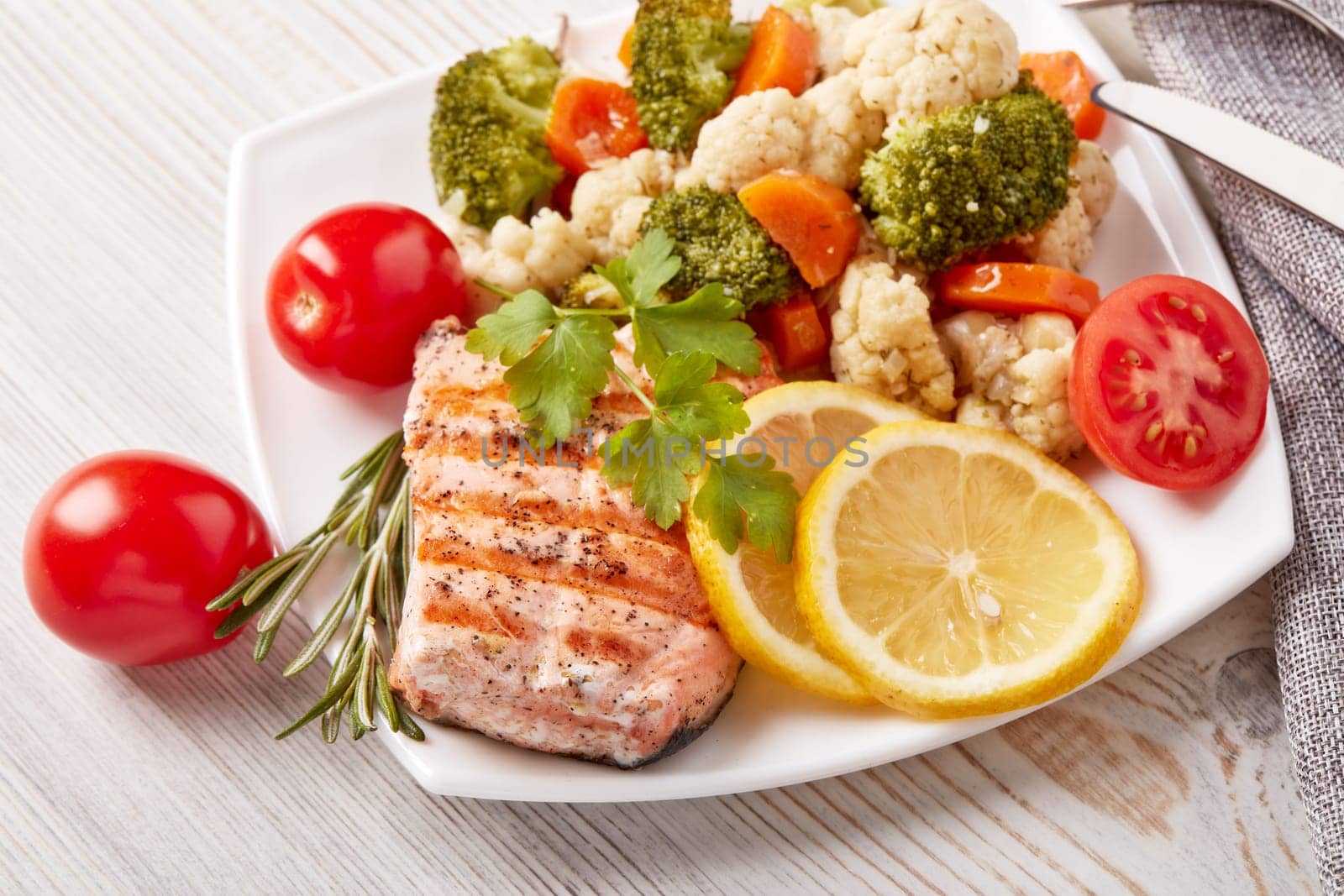 Grilled salmon fillet  garnished with vegetables, lemon, tomato, herbs lying on plate on wooden background. Top view. Healthy food. Keto diet.