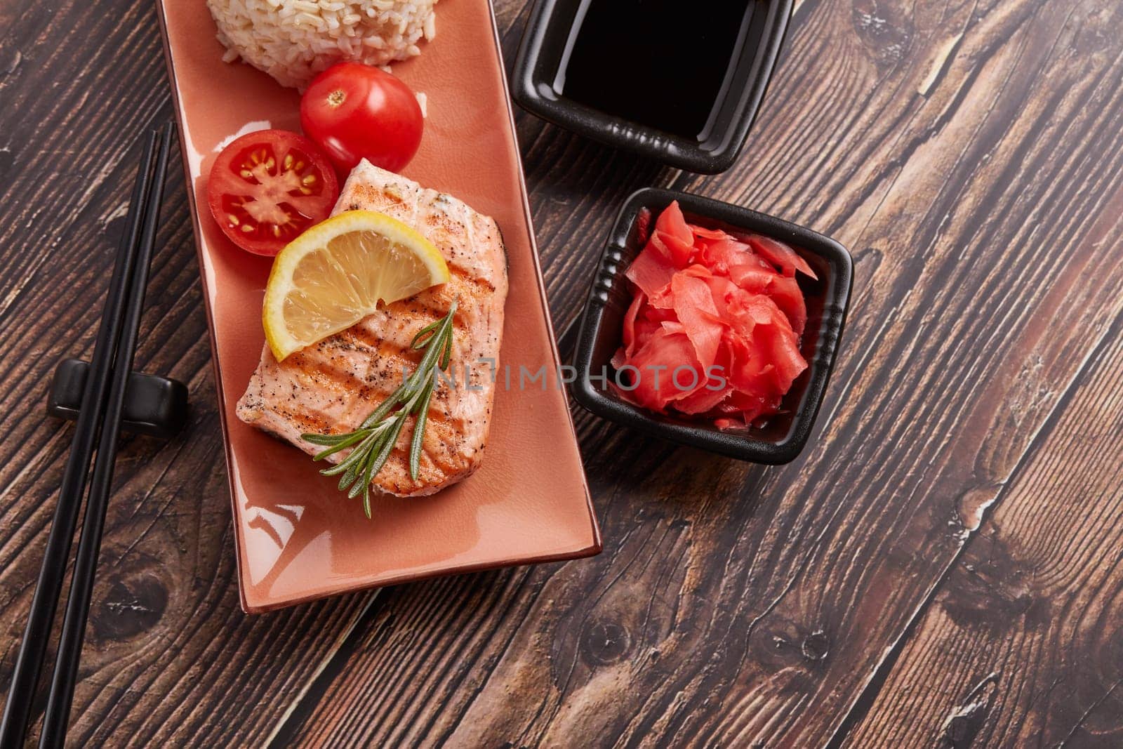 Grilled salmon fillet with rice, pickled ginger, soy sauce, tomato, lemon and herbs on dark wooden background. Top view. Healthy food.