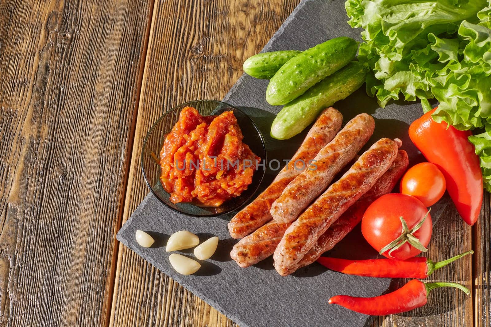 Grilled pork sausages  with fresh vegetables  on stone cutting board on wooden background. Top view