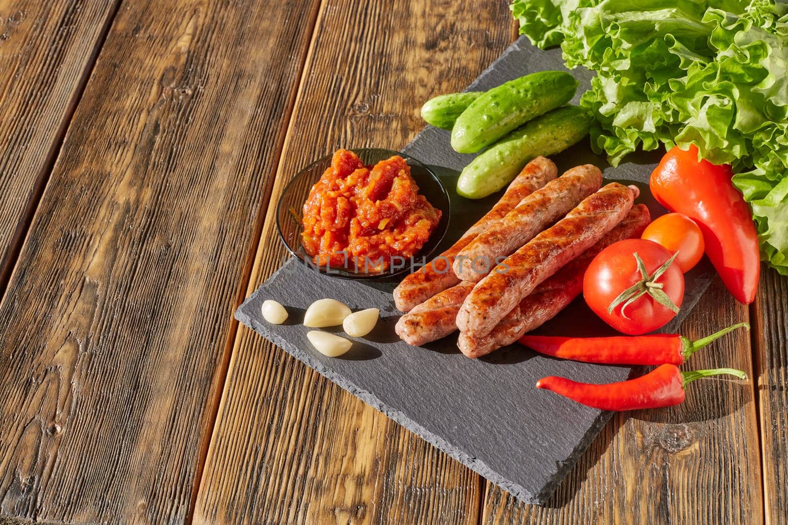 Grilled pork sausages  with fresh vegetables  on stone cutting board on wooden background. Top view with copy space.