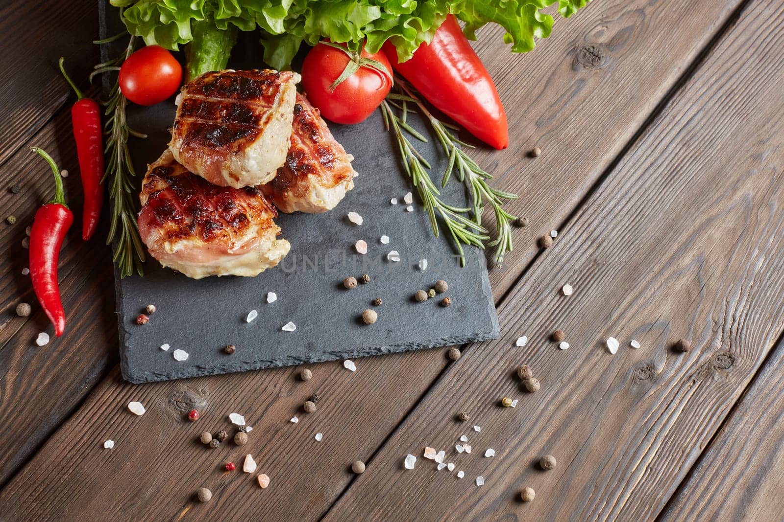 Bacon-wrapped grilled chicken (turkey)  fillet with fresh vegetables and spices on stone cutting board on wooden background. Top view with copy space