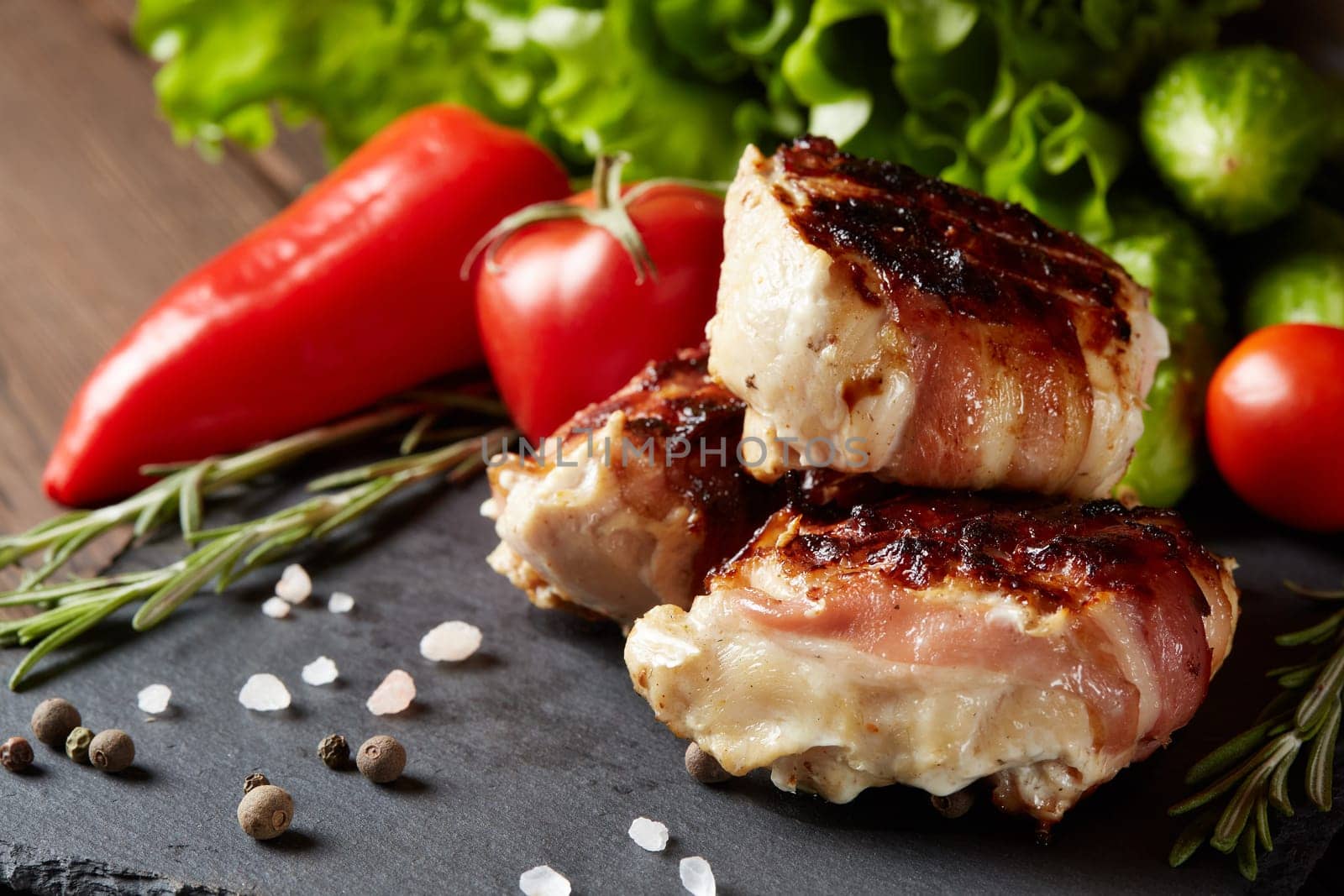 Bacon-wrapped grilled chicken (turkey) fillet with fresh vegetables and spices on stone cutting board on wooden background