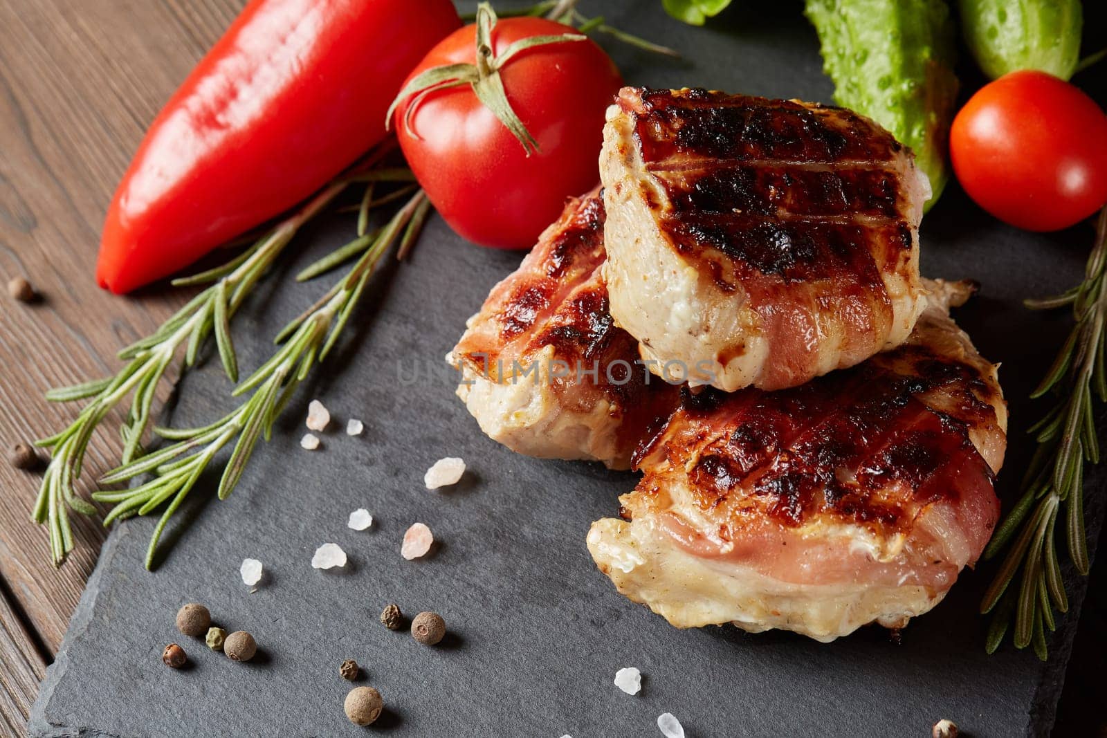 Bacon-wrapped grilled chicken (turkey) fillet with fresh vegetables and spices on stone cutting board on wooden background