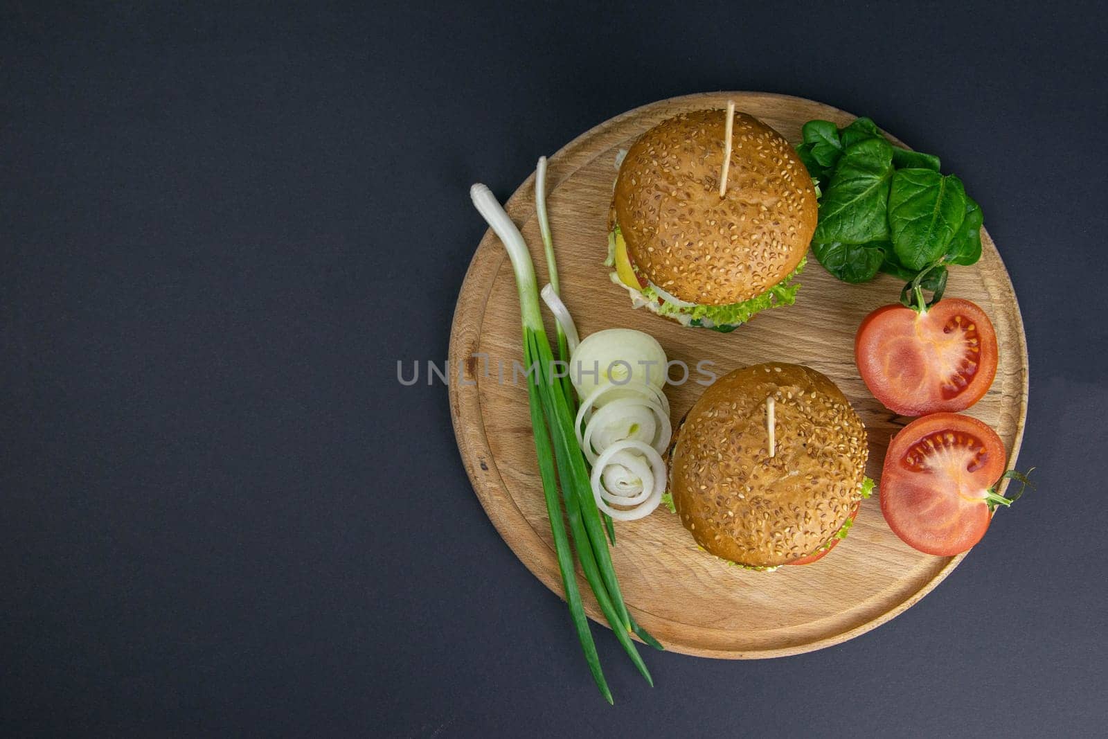 Two hamburgers close-up on a round wooden board, next to, tomatoes, chopped onions and spinach leaves. Top views with clear space