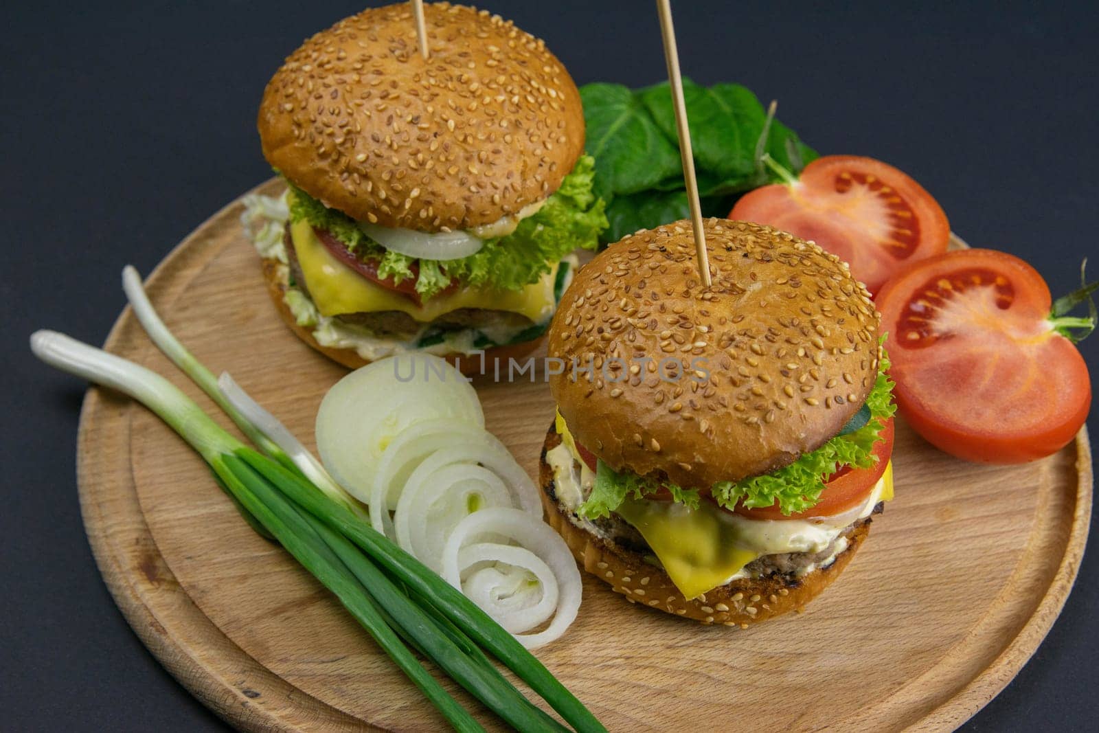 Two hamburgers close-up on a round wooden board, next to, tomatoes, chopped onions and spinach leaves.
