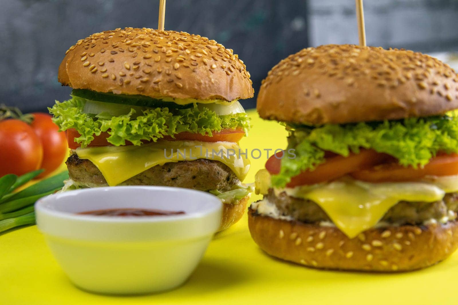 Two hamburgers close-up on a round wooden board, next to, tomatoes, chopped onions and spinach leaves.