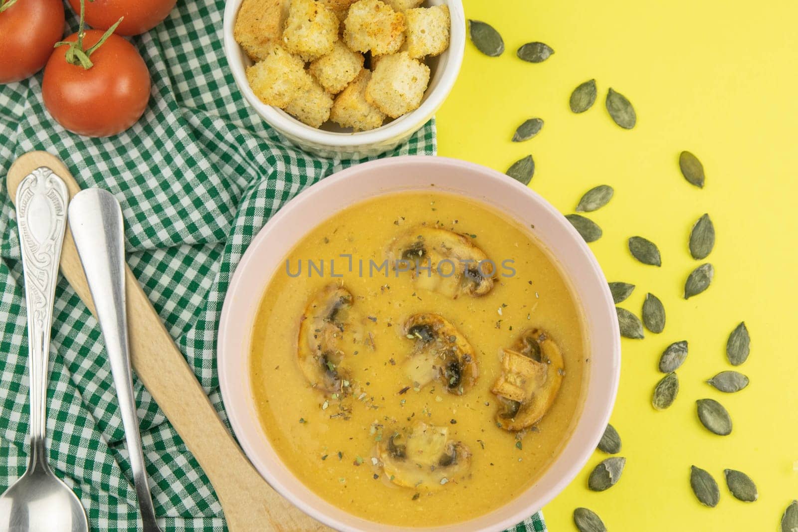 Pea soup with croutons stands on a green towel, next to there are tomatoes, toasts of pumpkin seeds, everything stands on the yellow table.