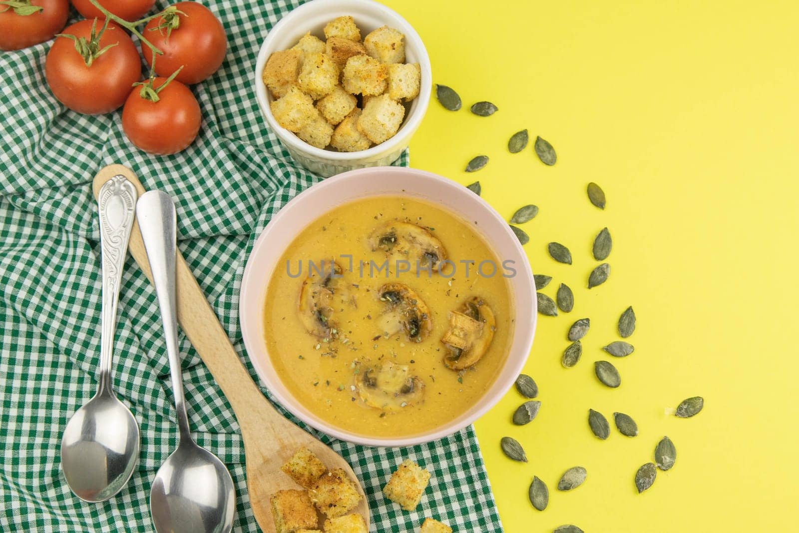 Pea soup with croutons stands on a green towel, next to there are tomatoes, toasts of pumpkin seeds, everything stands on the yellow table.