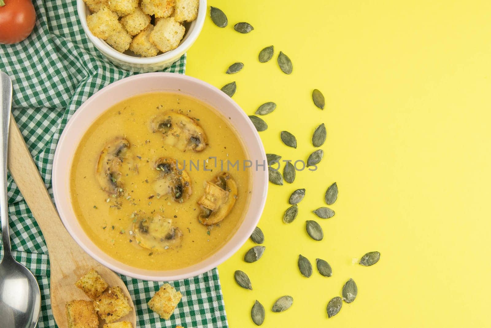 Pea soup with croutons stands on a green towel, next to there are tomatoes, toasts of pumpkin seeds, everything stands on the yellow table.