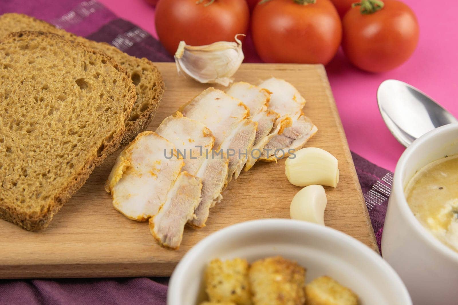Pea soup with toasts and pumpkin seeds, next to the wooden board sliced bacon, garlic and black bread, next to tomatoes.