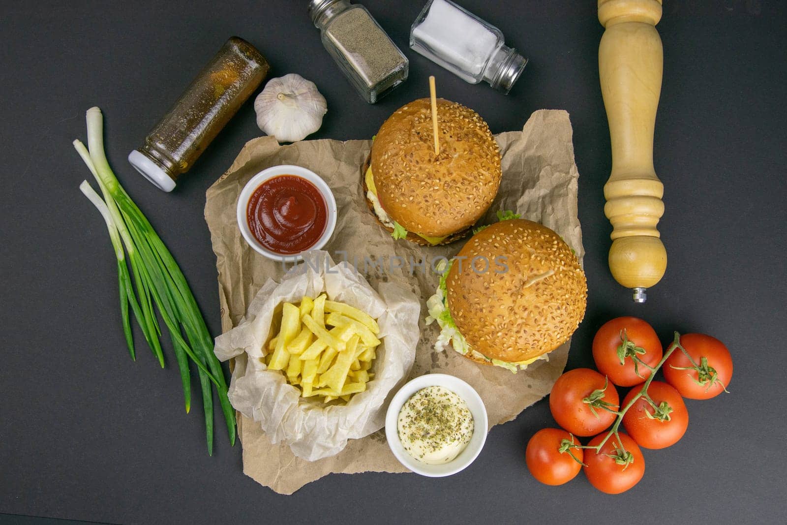 Close-up hamburger, sauces, french fries and tomatoes stand nearby, grill rack in the background.