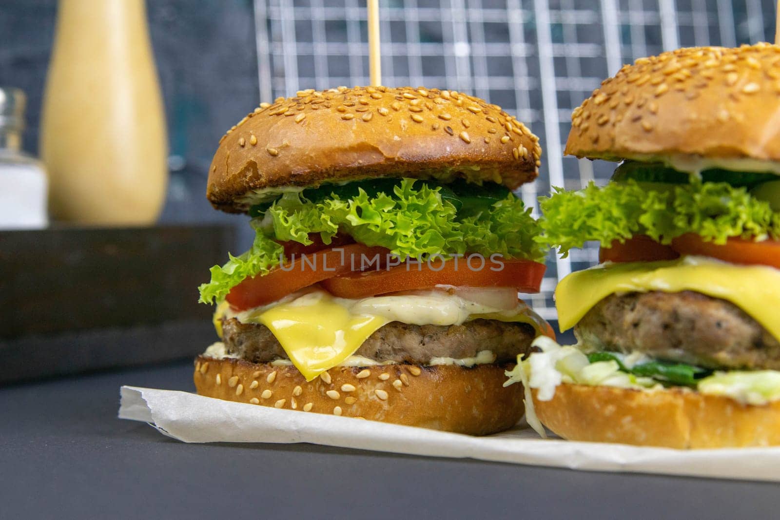 Close-up hamburger, sauces, french fries and tomatoes stand nearby, grill rack in the background.