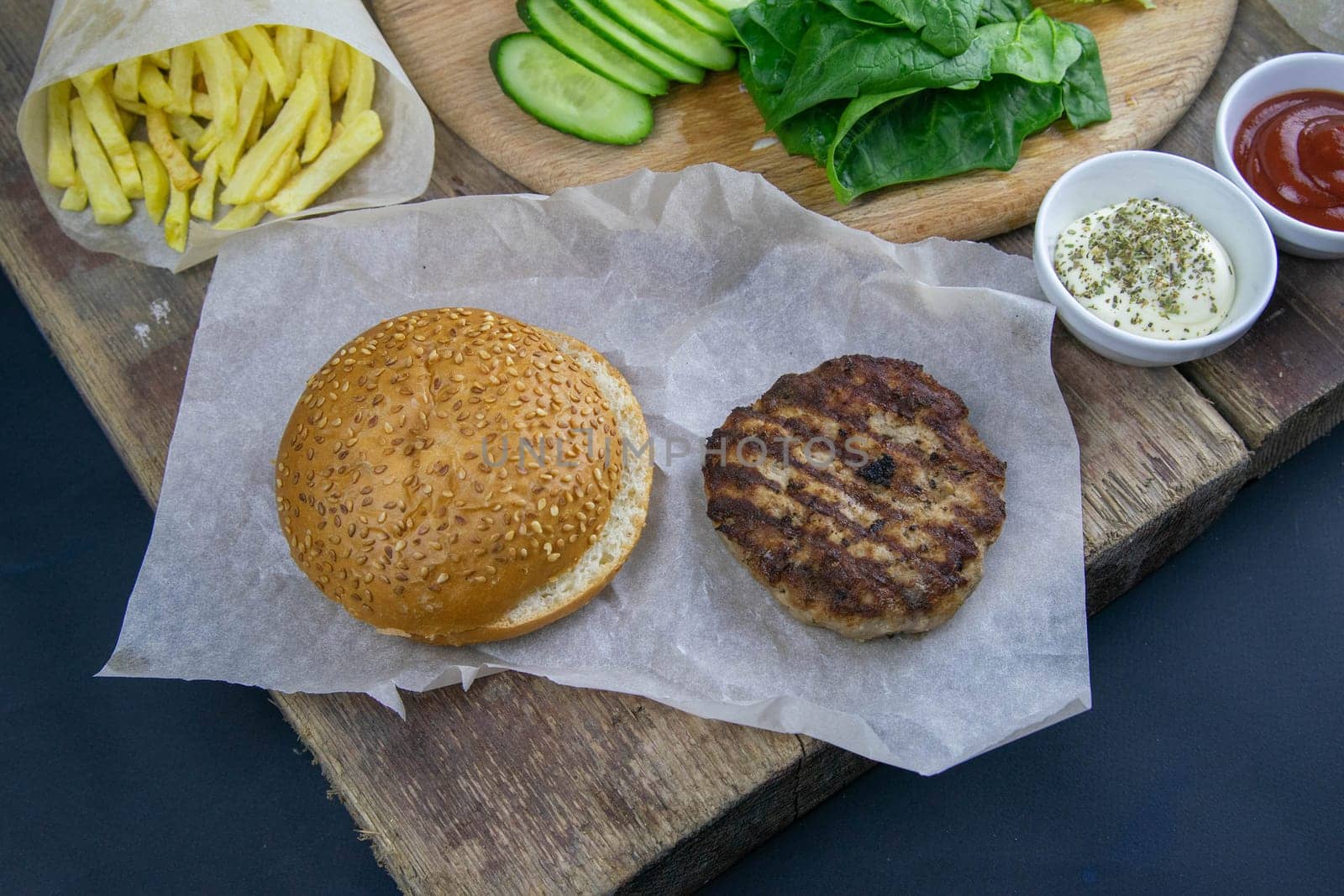 Preparing for the preparation of hamburgers. On a wooden board are lettuce leaves, chopped: tomatoes, cucumbers, onions, cutlets and sauces.