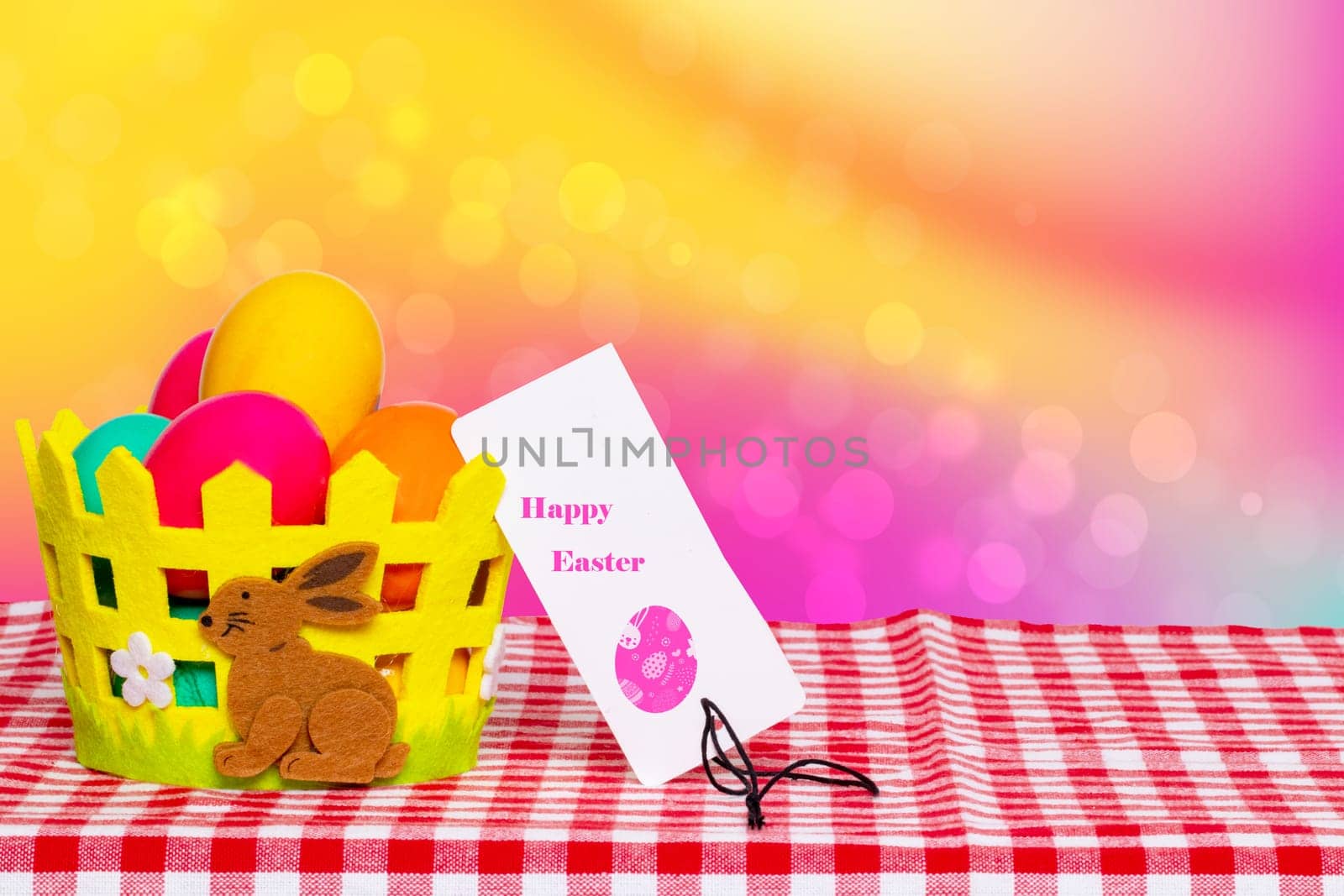 Table top on easter background. Colorful easter eggs in a basket with a bunny and a easter tag on picnic tablecloth over abstract rainbow backdrop. For your food and product display montage. Space.
