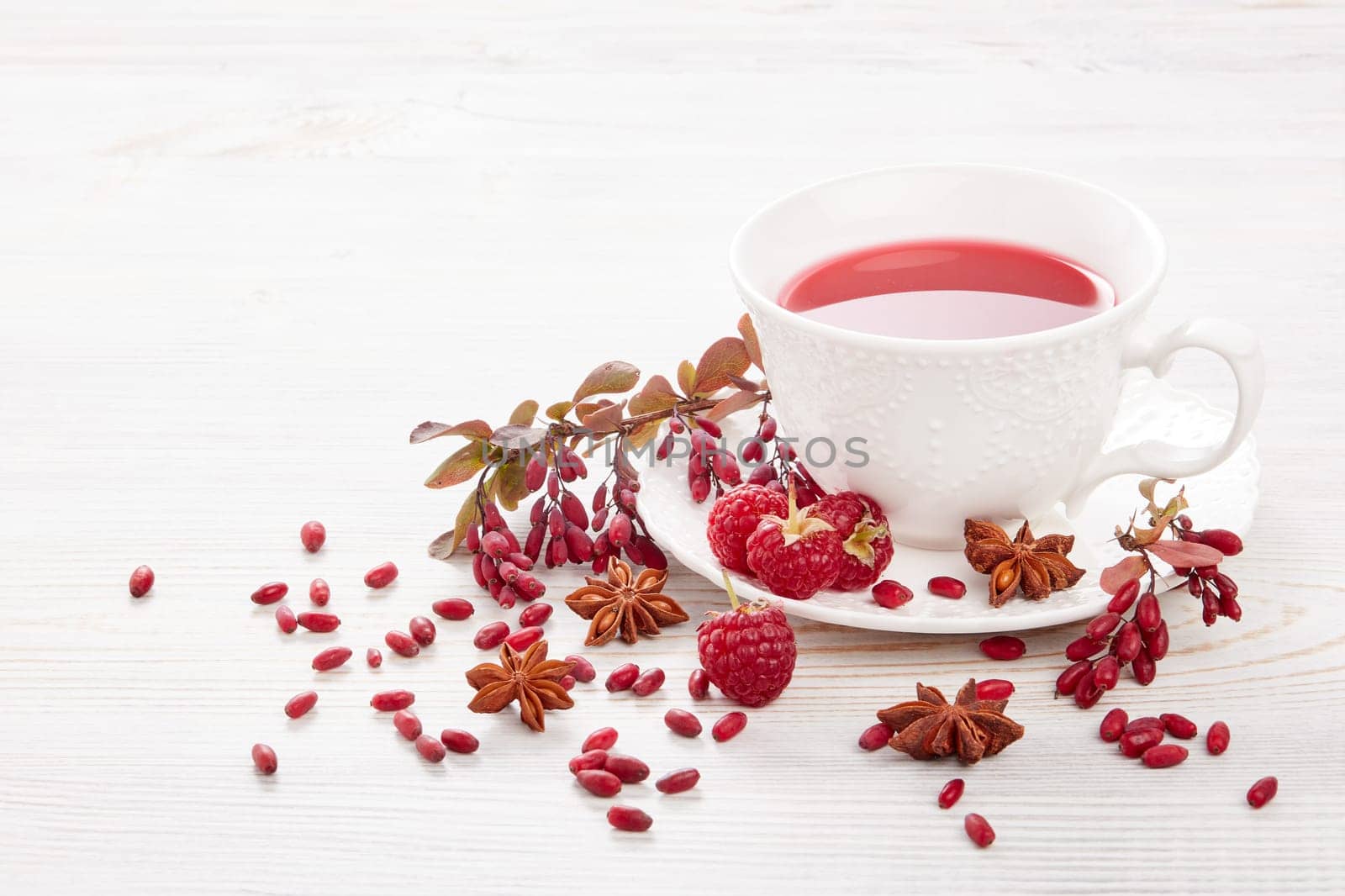 Healthy fruit tea with anise, barberries and raspberries in white porcelain cup and branch of berberry on light wooden background.