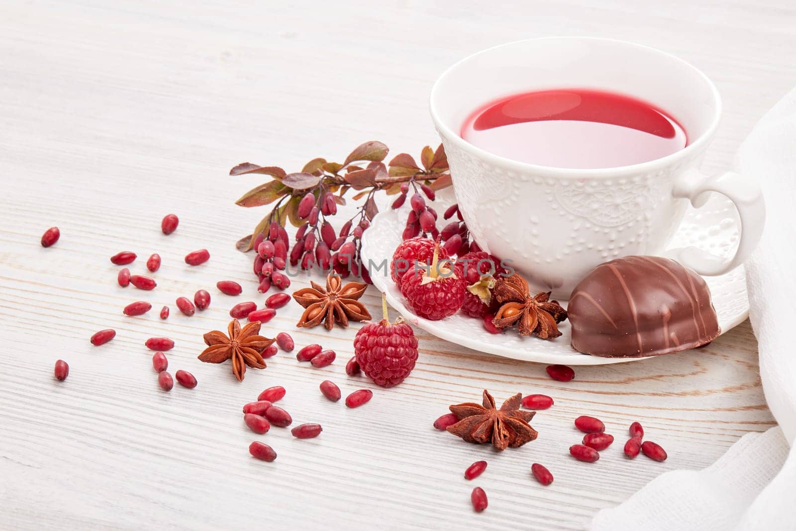 Healthy fruit tea with anise, barberries and raspberries in white porcelain cup and branch of berberry on light wooden background.