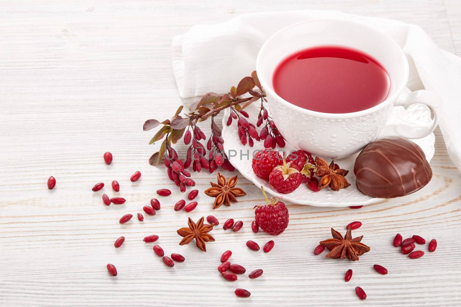 Healthy fruit tea with anise, barberries and raspberries in white porcelain cup and branch of berberry on light wooden background.