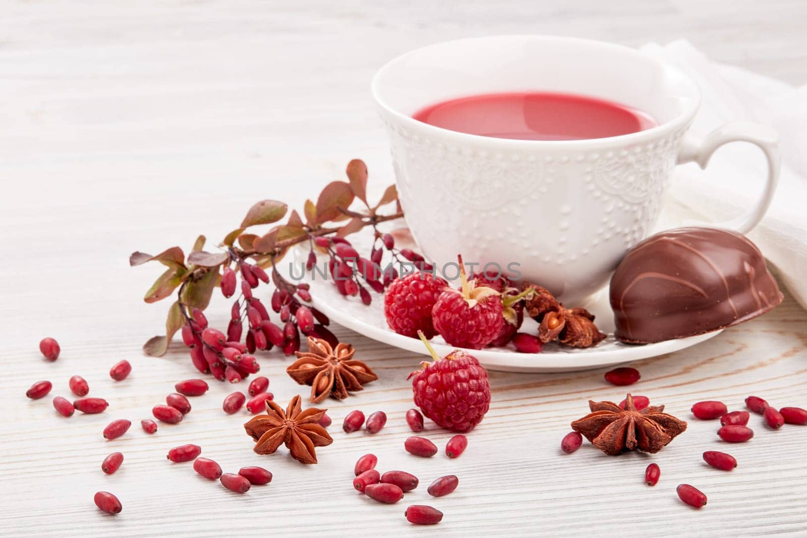 Healthy fruit tea with anise, barberries and raspberries in white porcelain cup and branch of berberry on light wooden background.