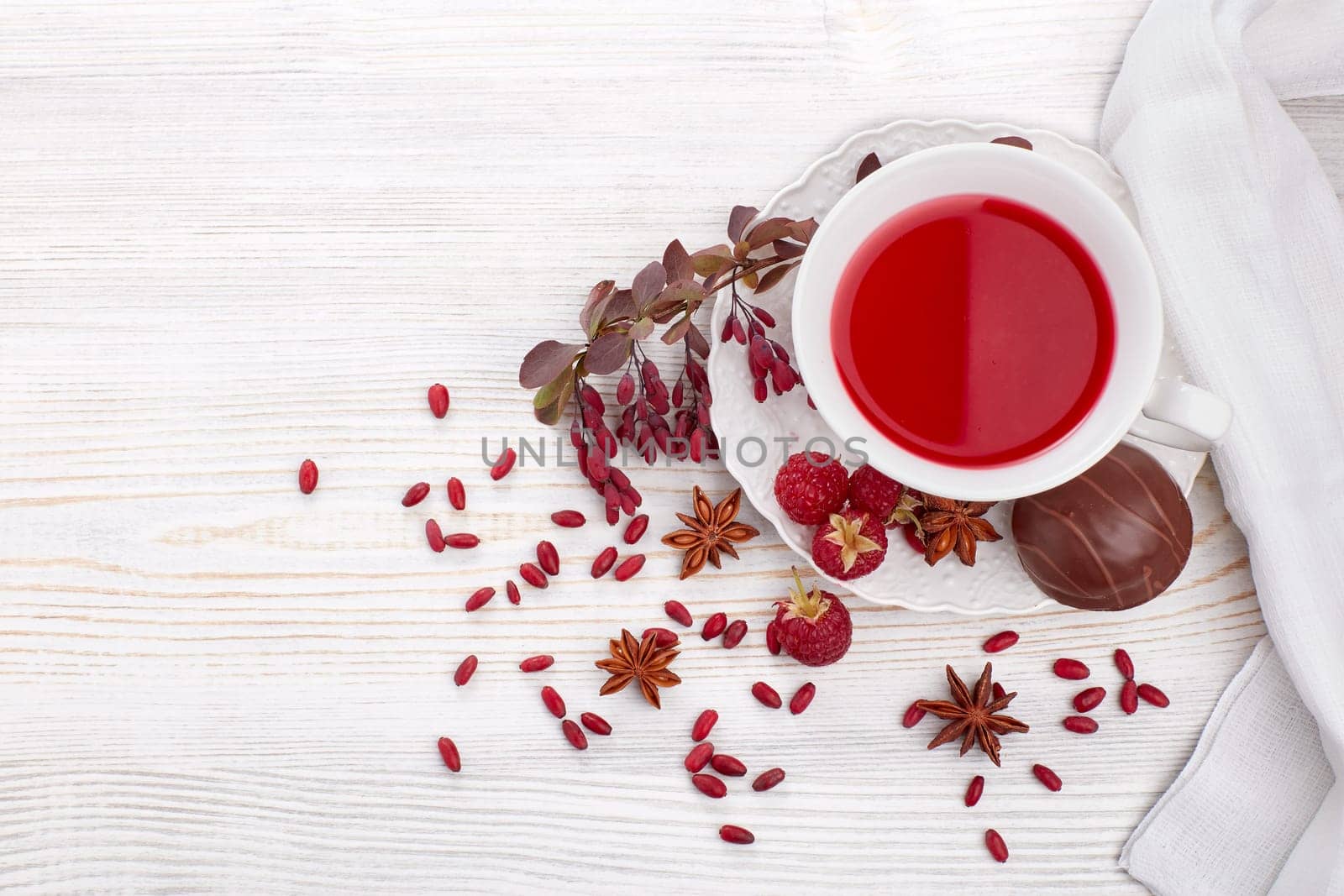 Healthy fruit tea with anise, barberries and raspberries in white porcelain cup and branch of berberry on light wooden background. Top view