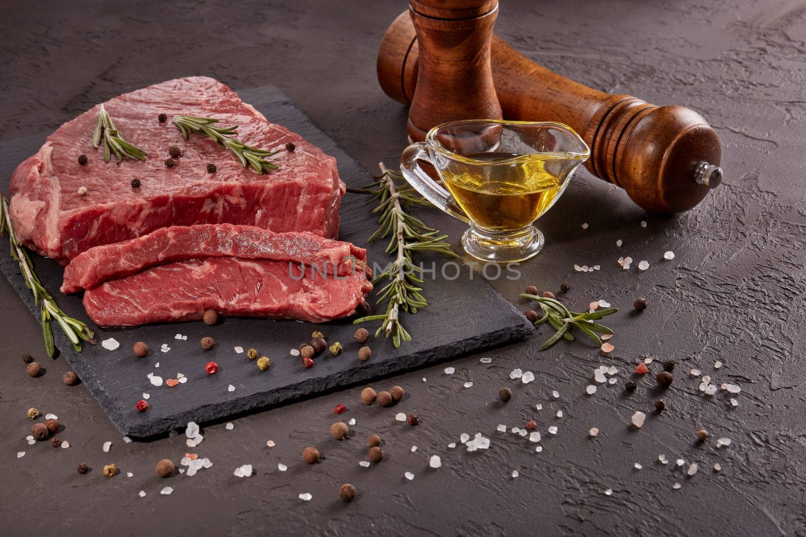 Still life with raw meat beef fillet on black stone cutting board,  rosemary twigs, set of different peppers, salt and olive oil on dark background. Fresh beef piece for steak or grilled barbecue