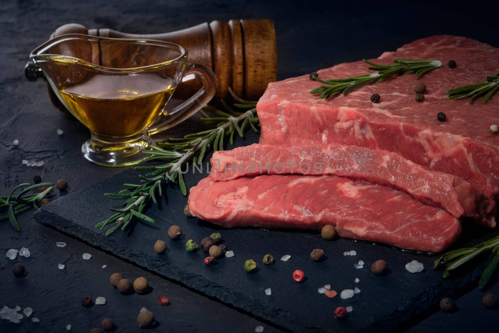 Still life with raw meat beef fillet on black stone cutting board,  rosemary twigs, set of different peppers, salt and olive oil on dark background. Fresh beef piece for steak or grilled barbecue