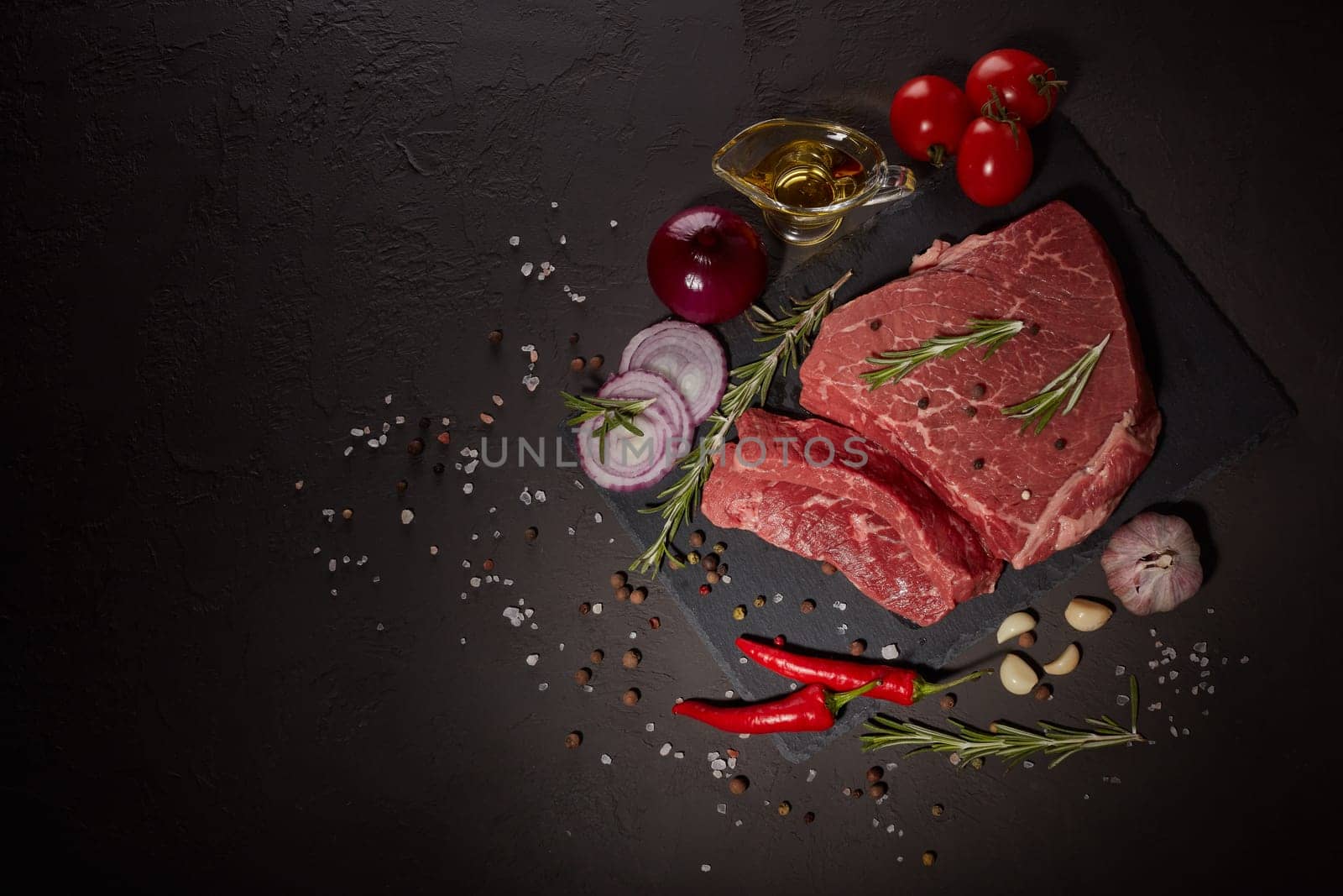 Raw meat beef fillet on stone cutting board, rosemary, set of different peppers, vegatables, olive oil on dark background. Fresh beef piece for steak or grilled barbecue. Top view with copy space