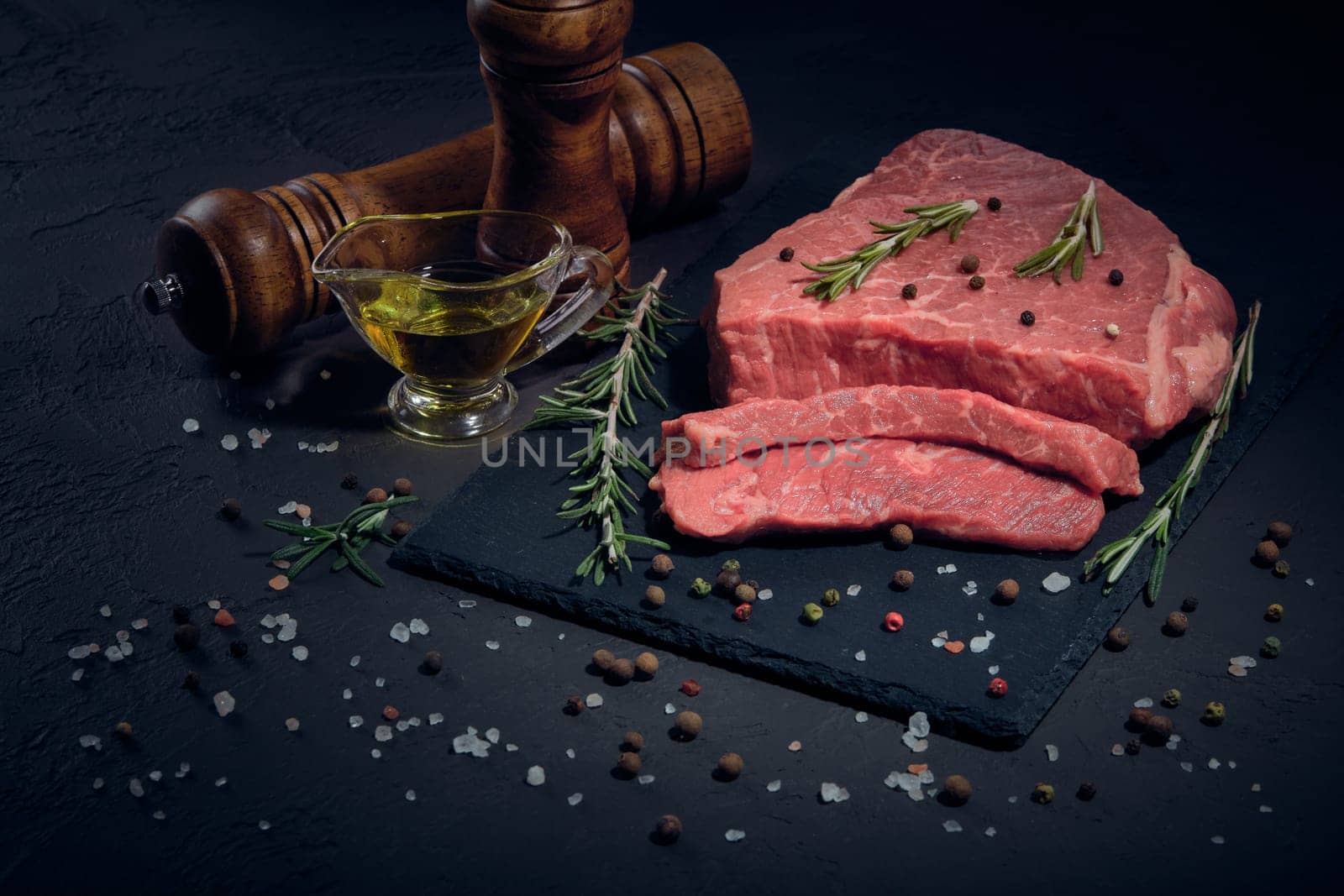 Still life with raw meat beef fillet on black stone cutting board,  rosemary twigs, set of different peppers, salt and olive oil on dark background. Fresh beef piece for steak or grilled barbecue