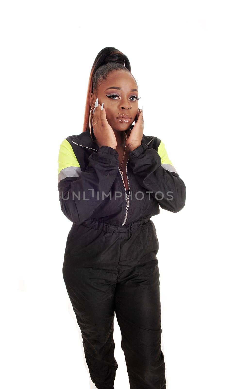 A gorgeous young African American woman standing in the studio in.a black track suit, her hands on her face, isolated for white background.