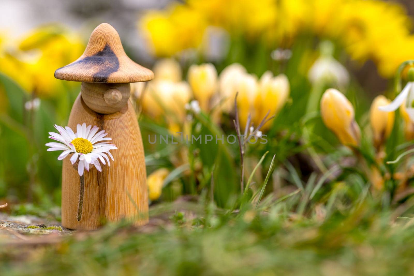 Wooden elf figure with a daisy in hand is standing in a flower bed in front of blurred yellow flowers