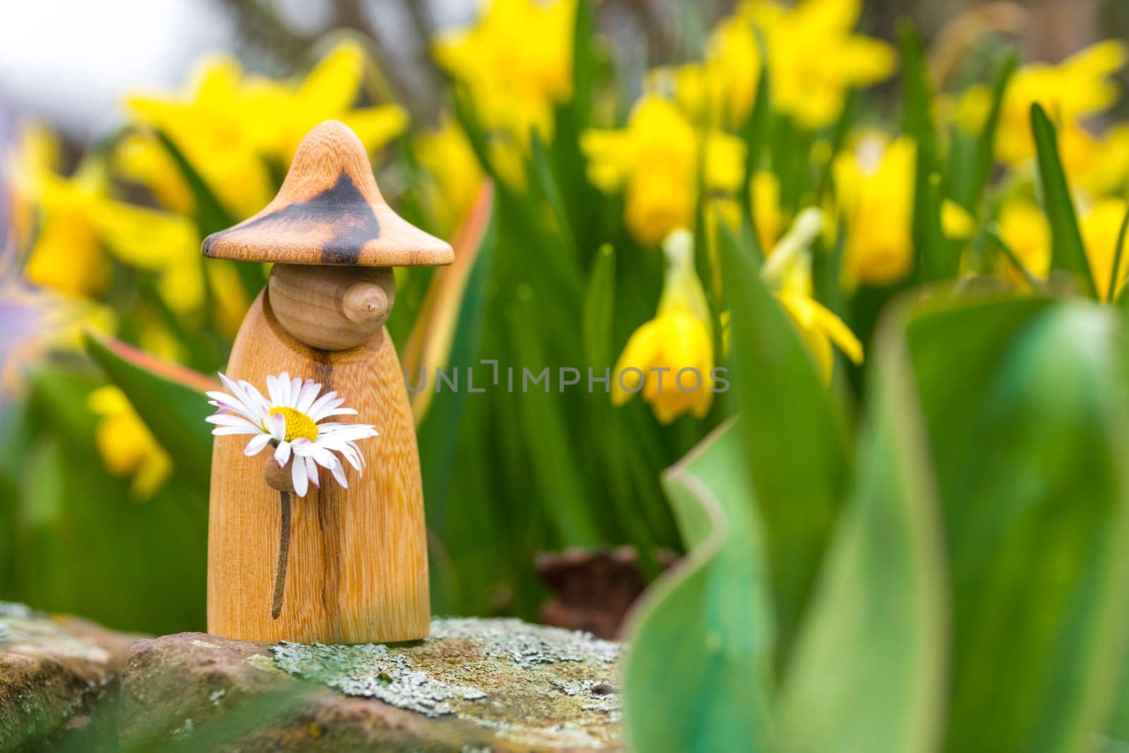 Wooden elf figure with a daisy in hand is standing in a flower bed in front of blurred yellow flowers