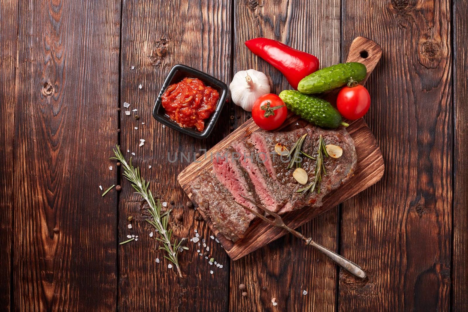 Grilled meat. Medium rare beef steak on wooden cutting board, rosemary twigs, spices and vegatables on brown wooden background. Top view. Steak menu