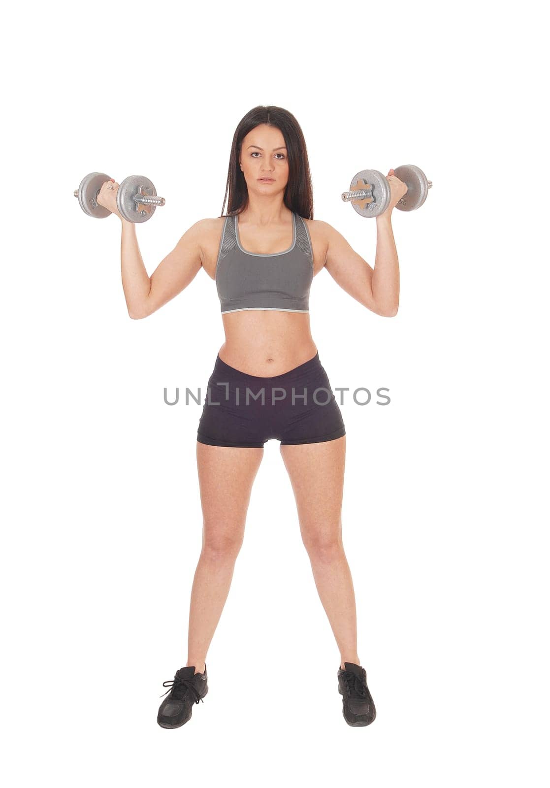 A beautiful young woman standing in shorts and sports bra from the.front lifting her two dumbbells, isolated for white background.