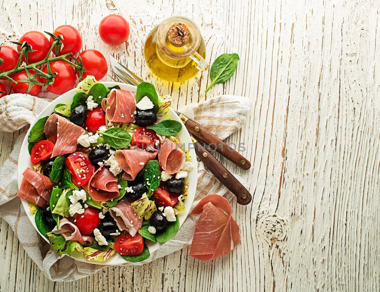Healthy green salad with prosciutto, olives, tomato and fresh cheese on wooden table background. Healthy meal.
