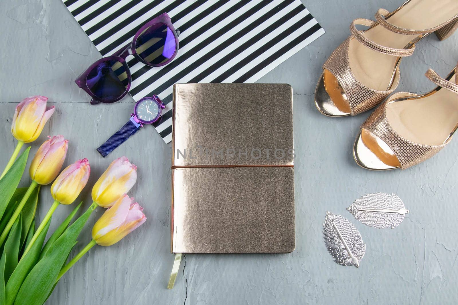 A gold notebook with women's shoes, tulips, glasses and a watch on a concrete background. Flet lay.