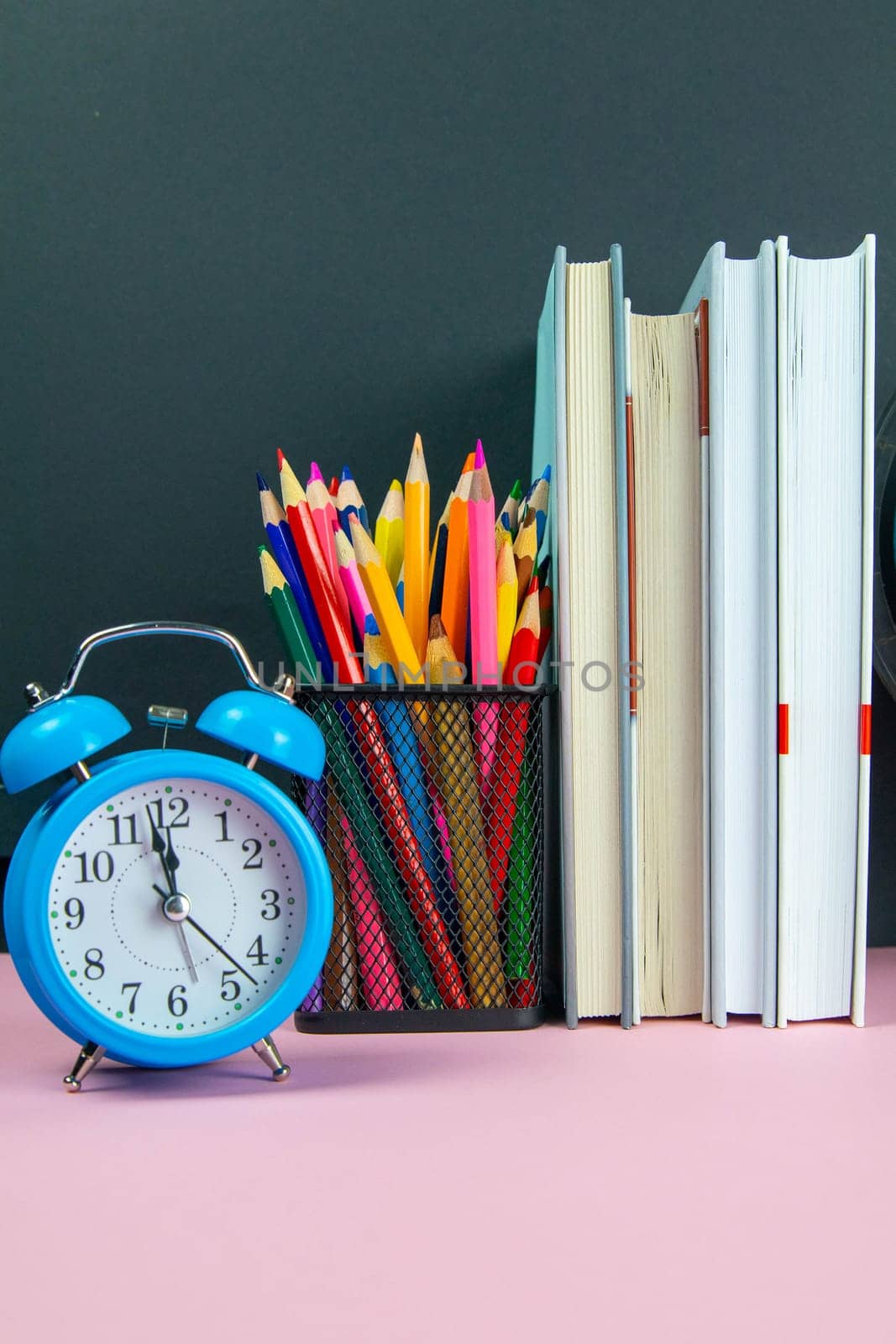 A stack of books, a clock and a glass with multi-colored pencils.