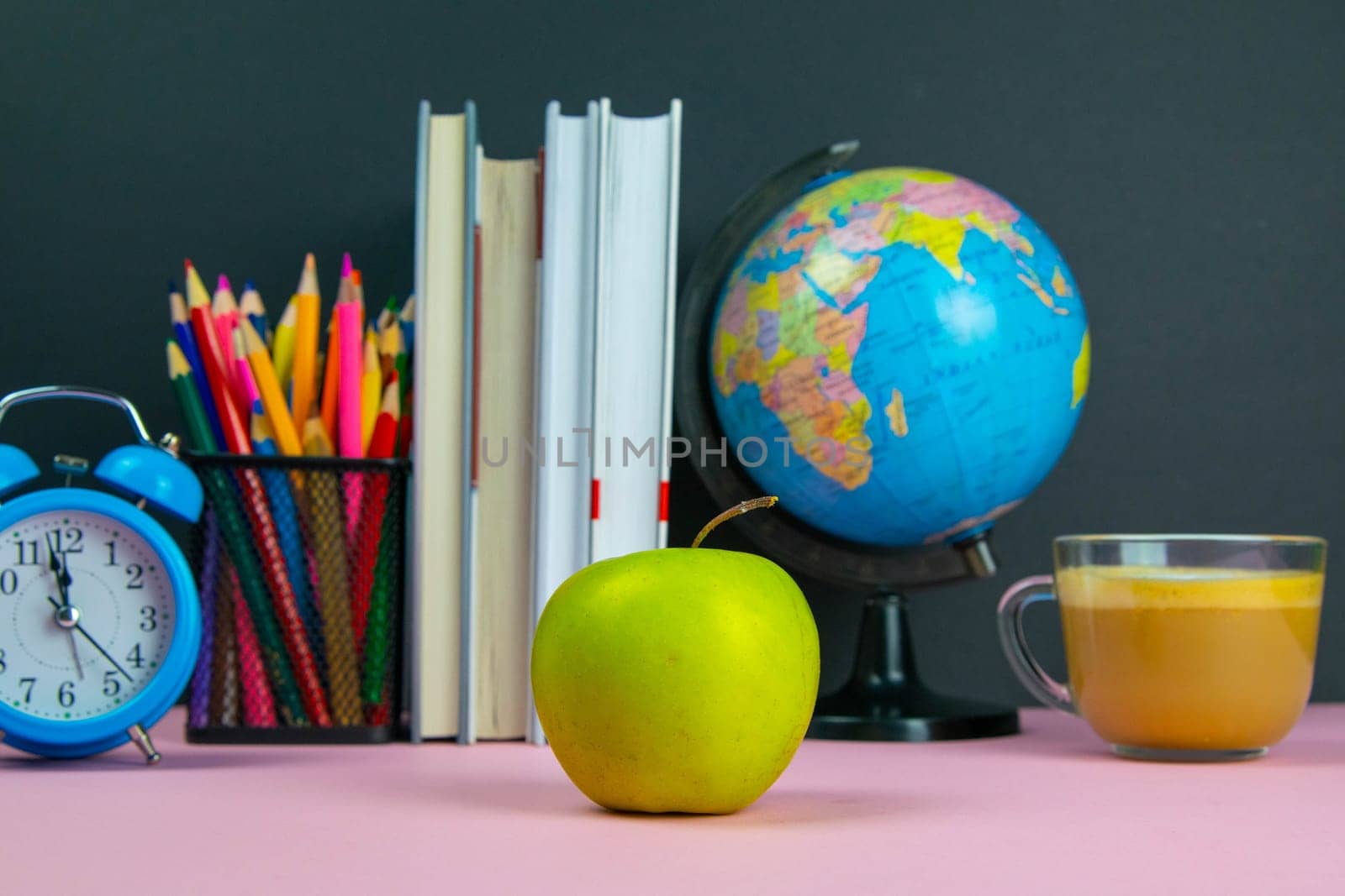 Apple is in the foreground and behind him are a stack of books, a globe, a clock and a glass of pencils.