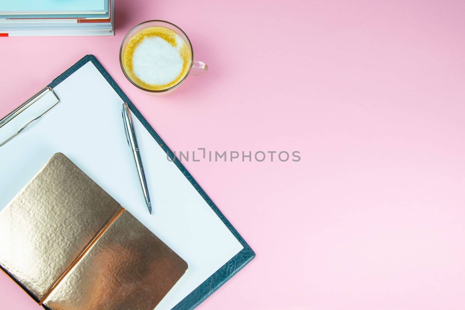 The pen and gold notebook stand on a tablet with a white sheet next to a glass of coffee.
