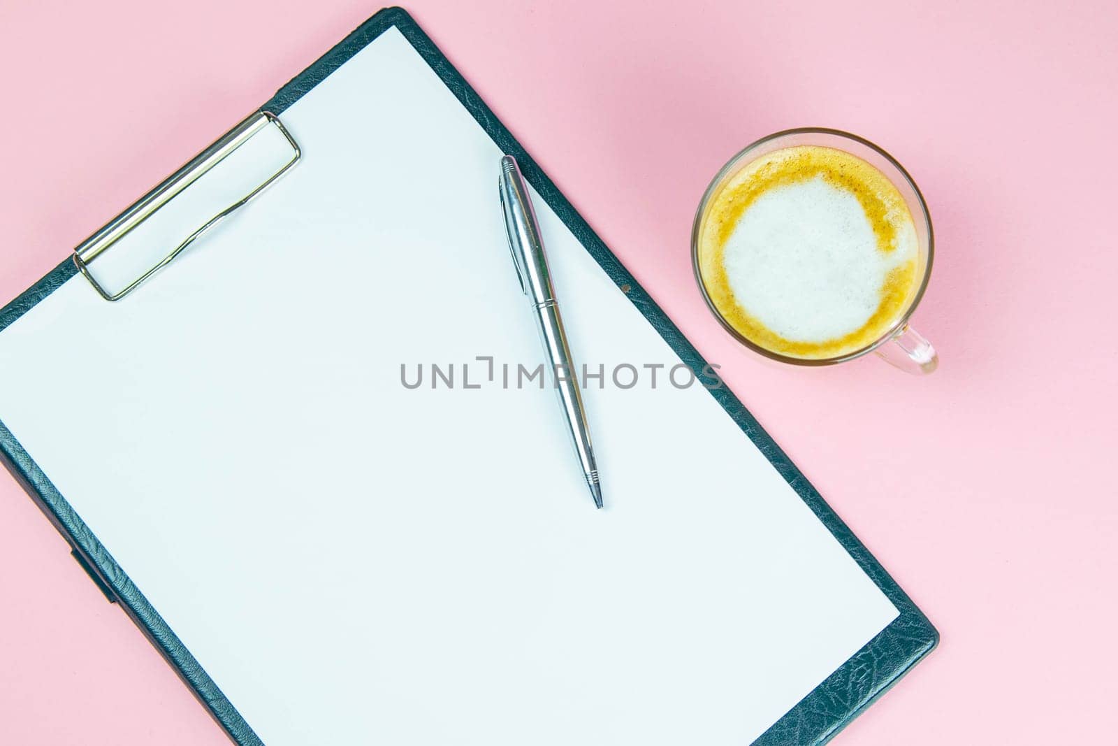 Tablet with a white sheet on which there is a silver pen and next to there is a glass with a coffe with milk.